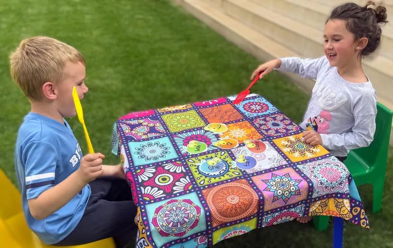 Two children at a colourful patterned table play a sound game with fly swatters, a girl laughing as she reaches across