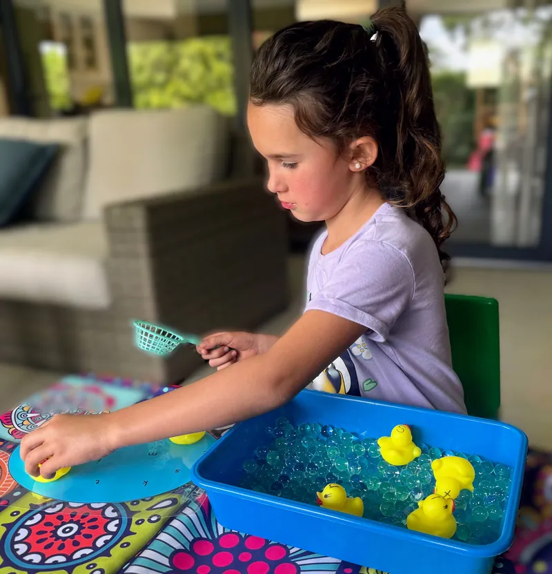 A girl scoops rubber ducks from a tub of water beads with a little net at a phonics sensory play station